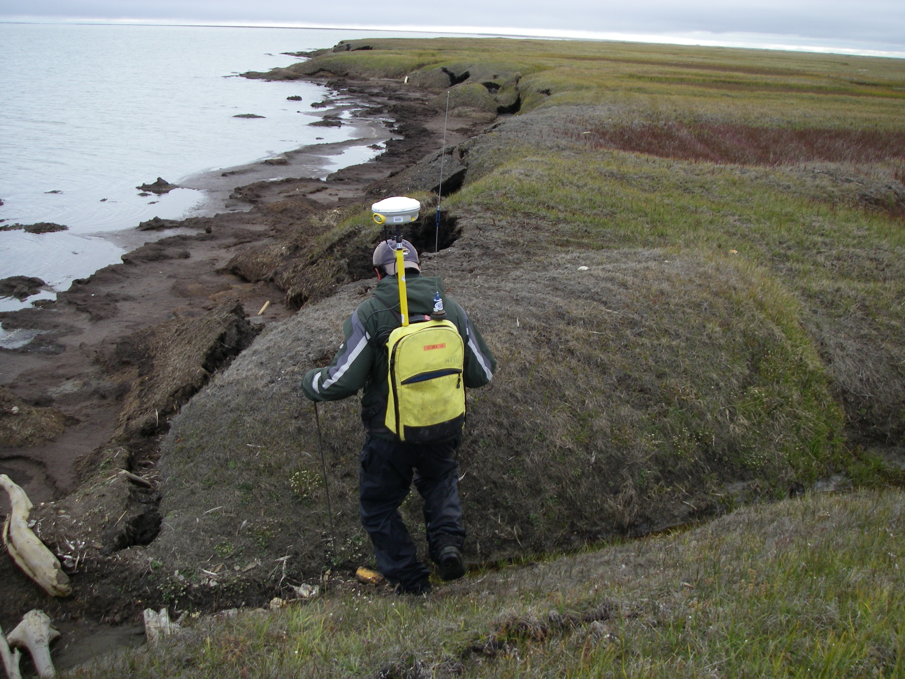 Image of a person walking along an irregular and eroded coastline with a backpack equipped with some technical instrumentation.
