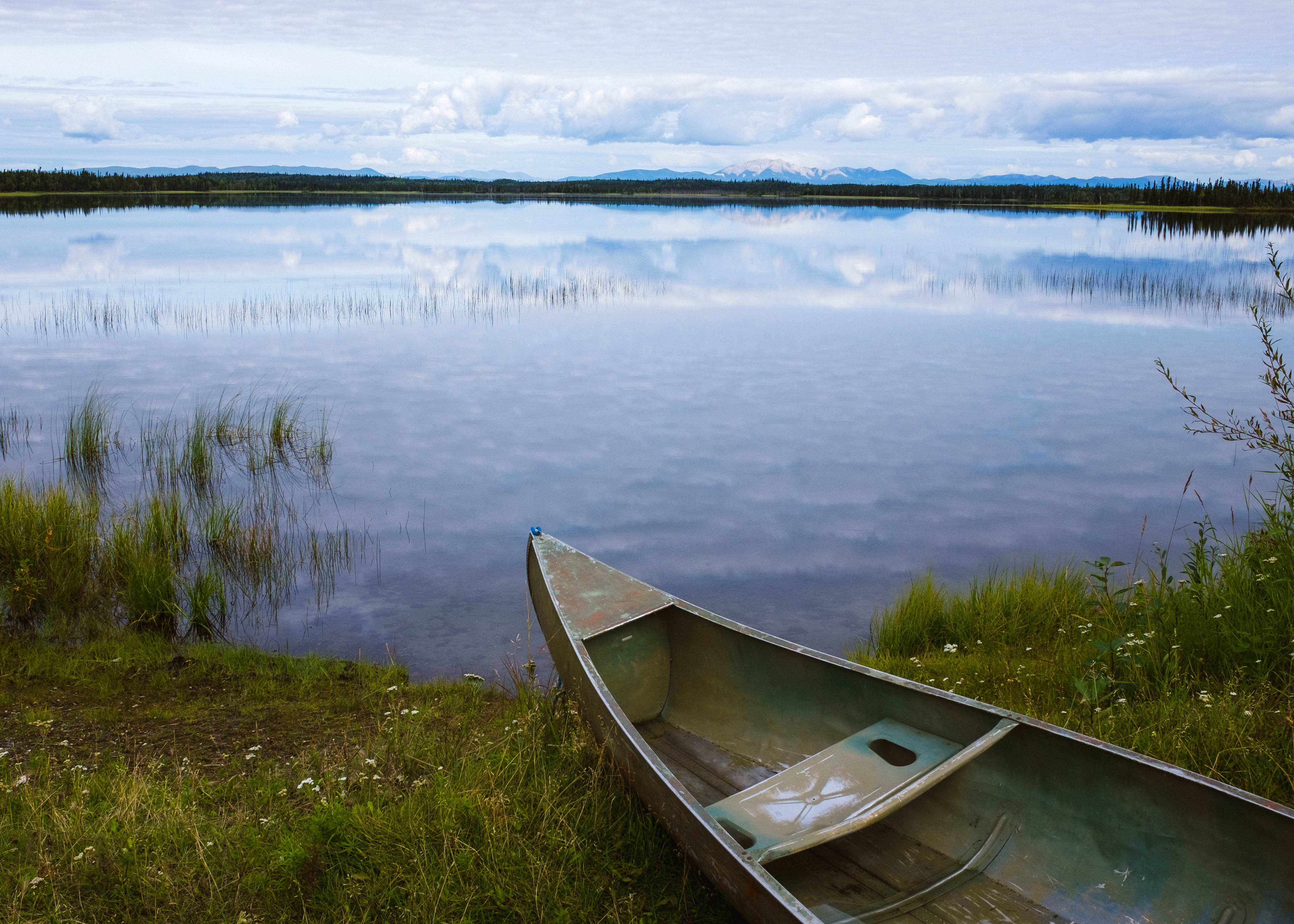 Image of a canoe sitting on the grassy bank of a calm lake with mountains in the distance.