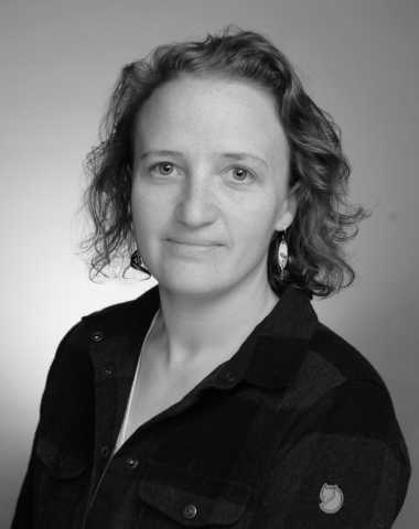 Black and white photo of a women in a flannel shirt. She is white with chin-length hair and earrings. 